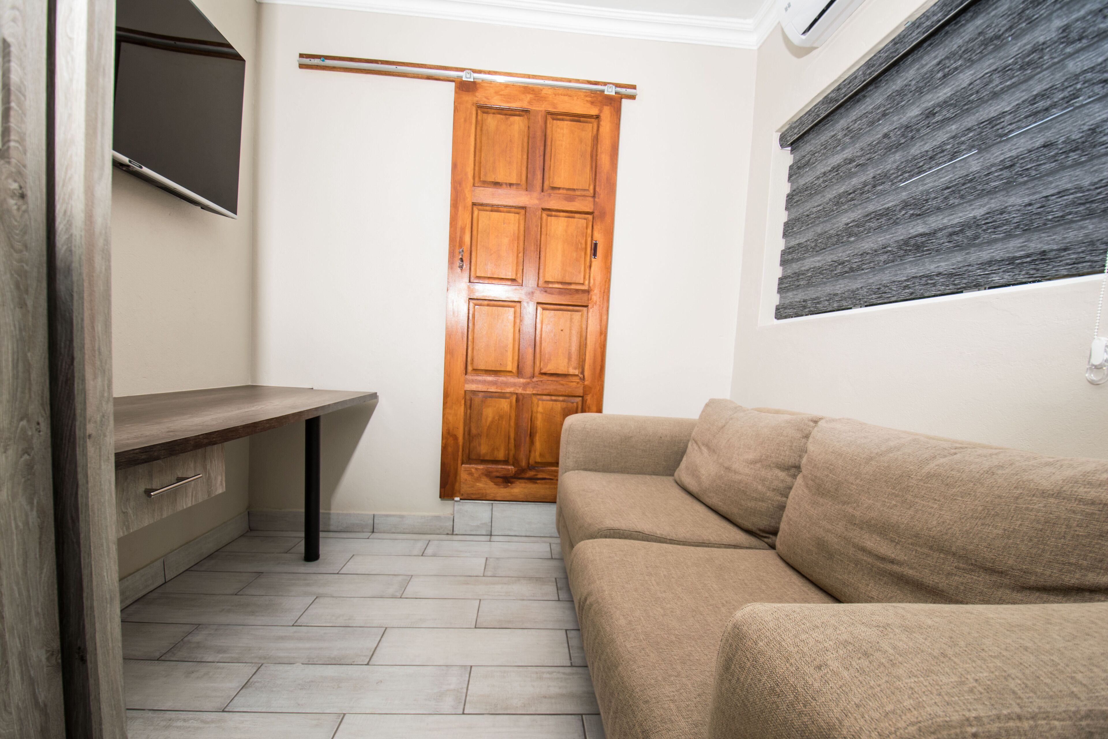 A room with a wooden door, a beige sofa, a wooden table, and a window with blinds.
