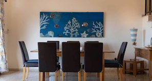 Un comedor con una mesa de madera y cuatro sillas, un cuadro azul con vida marina en la pared, y un pequeño aparador de madera con adornos.