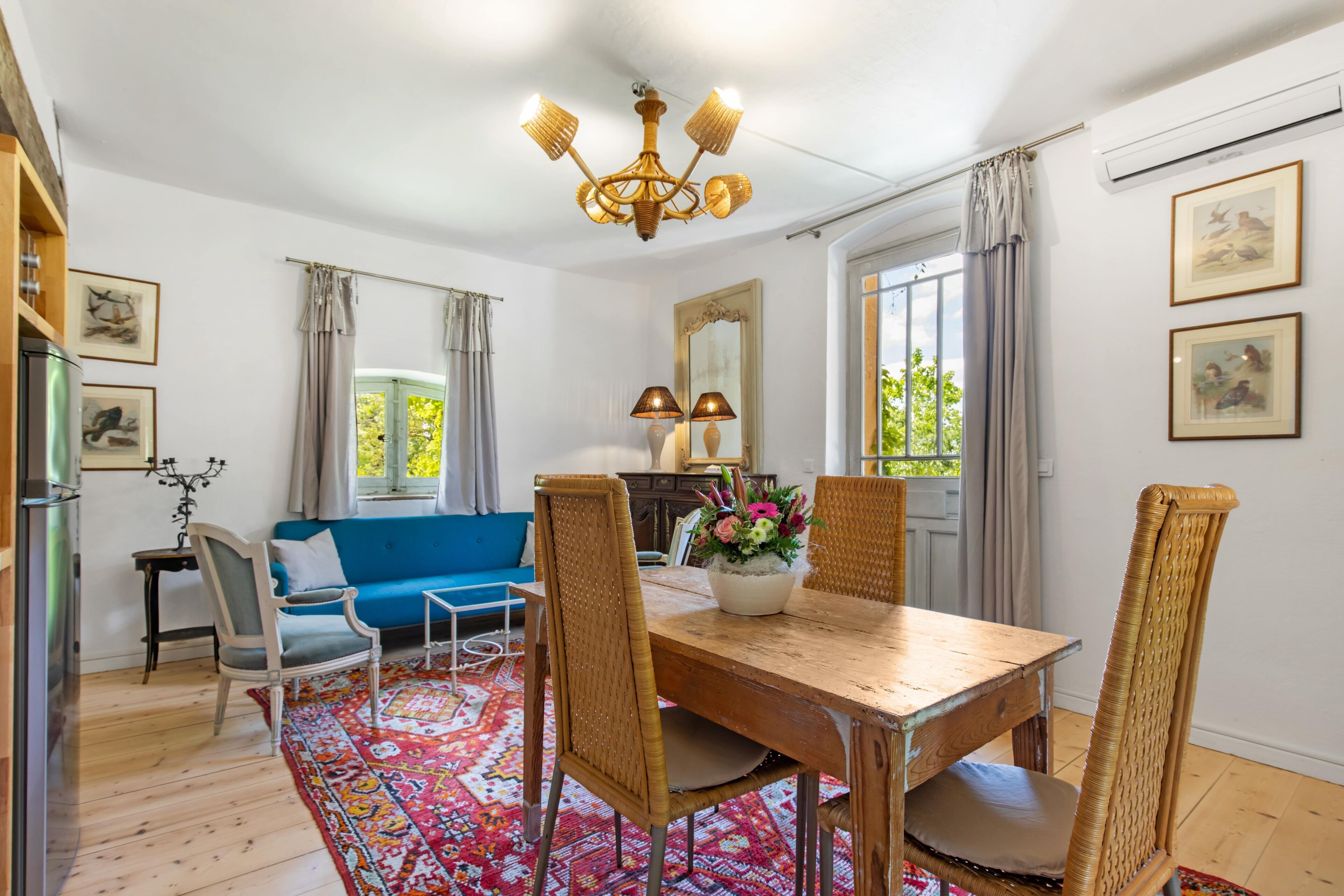 Un comedor con una mesa de madera, sillas de mimbre, un sofá azul y una araña.