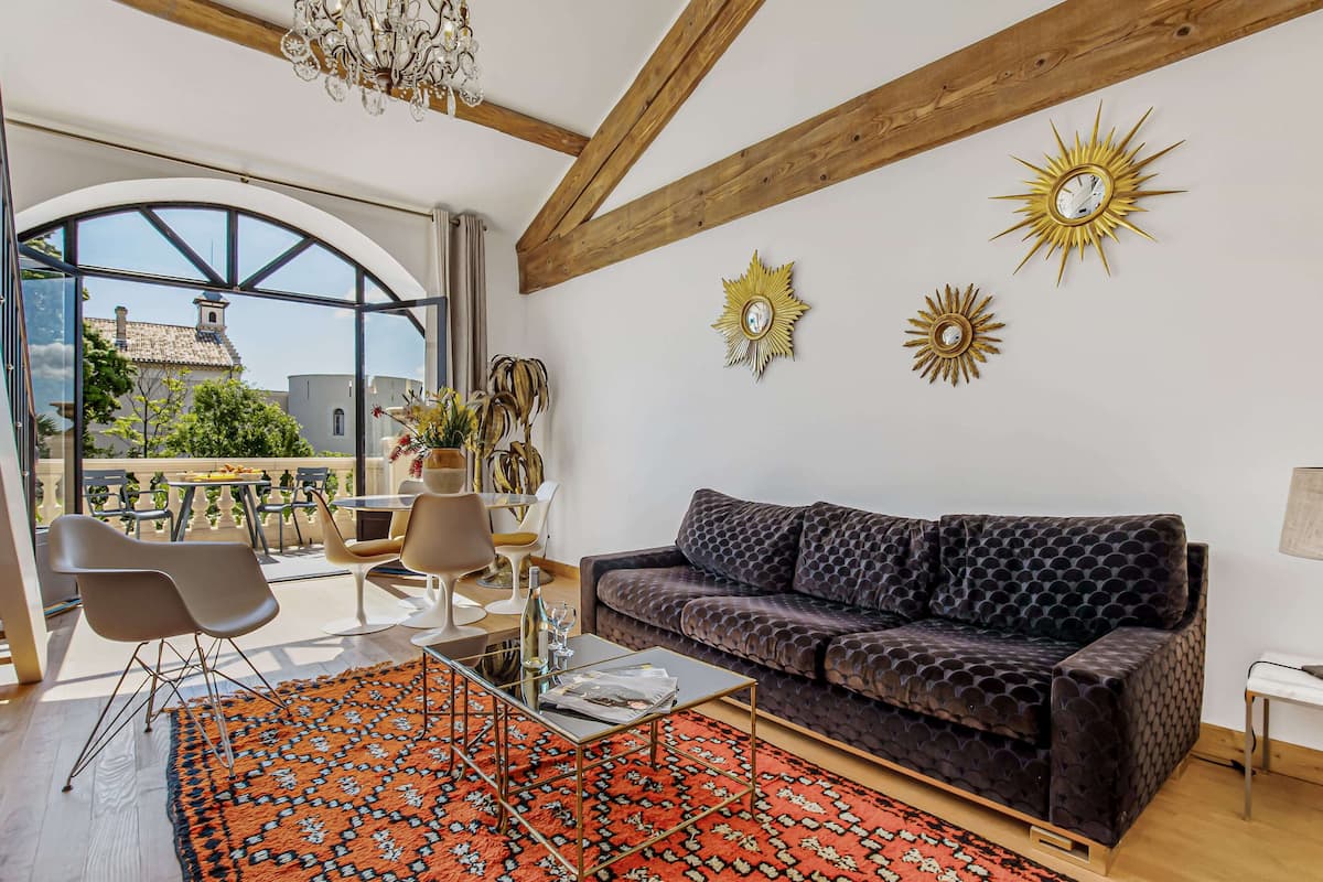 Una sala moderna con un sofá estampado oscuro, una mesa de centro de vidrio y una alfombra roja con diseño. Se observan dos adornos en forma de estrella en la pared y una viga de techo de madera.