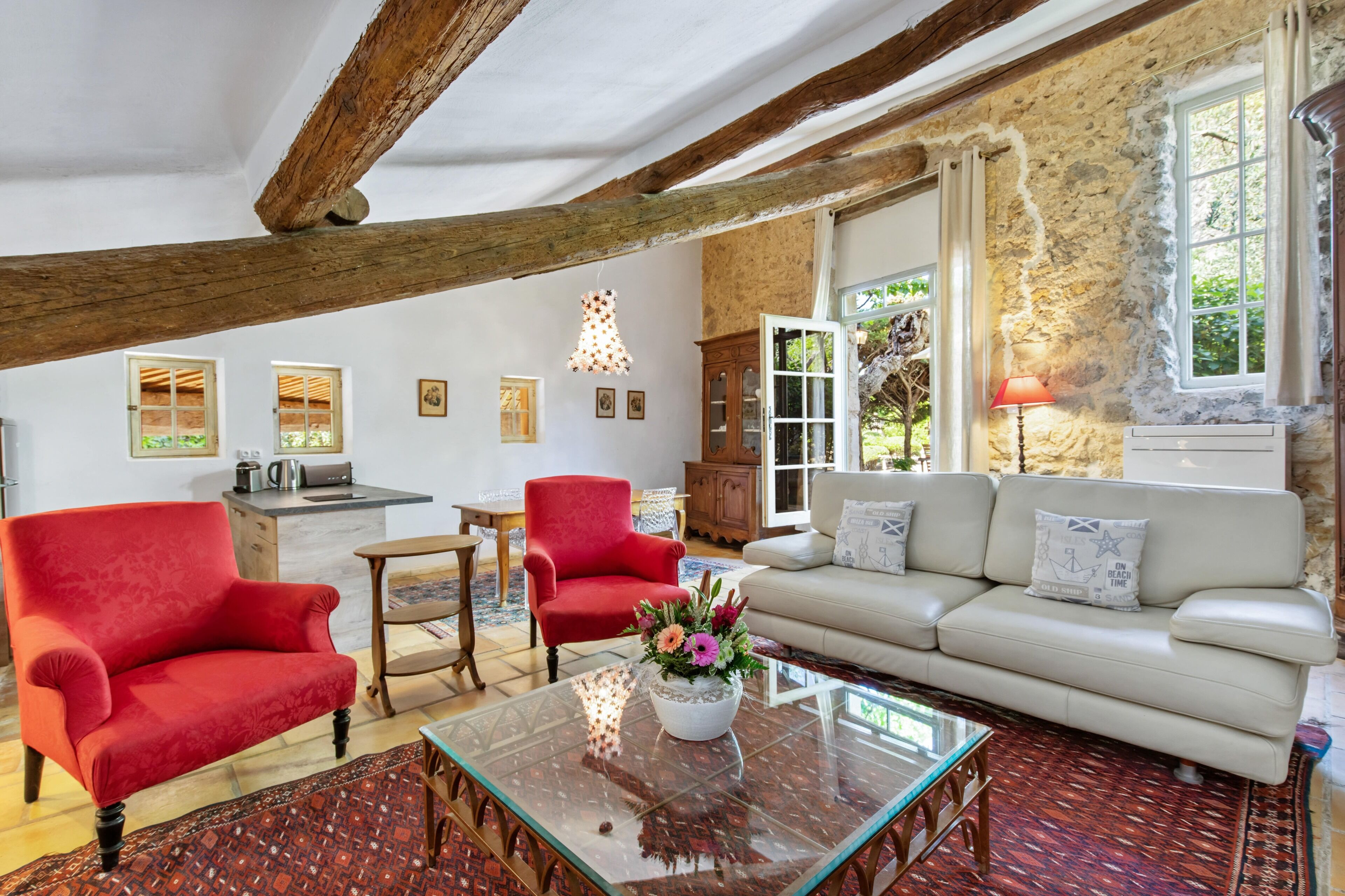 A spacious living room with a high ceiling, exposed wooden beams, a red armchair, a white sofa, and a glass coffee table with a floral centerpiece.