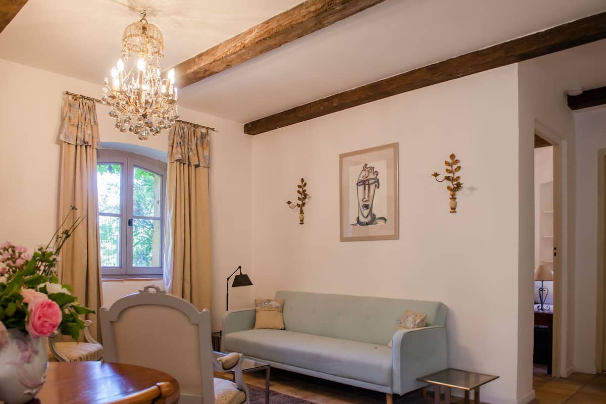 Una sala de estar con un sofá verde claro, una mesa de comedor de madera con sillas, una araña y una ventana con cortinas.