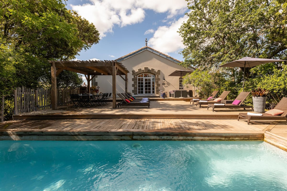 Un área de piscina con sillas de descanso y una pérgola, rodeada de árboles y un edificio con techo de tejas.