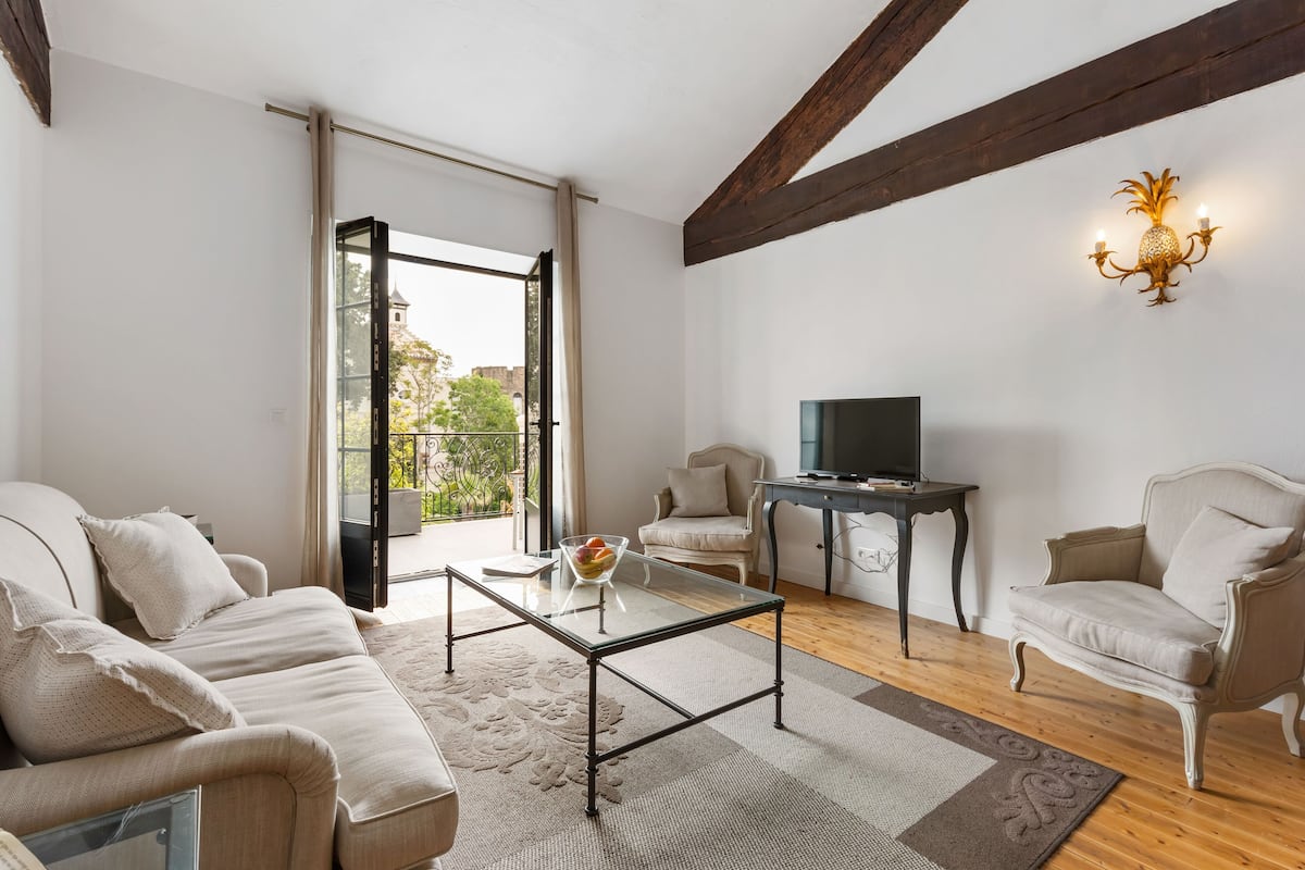 Una sala de estar moderna con una mesa de centro de vidrio, sofás color beige, un televisor en un mueble bajo y un balcón con vistas.