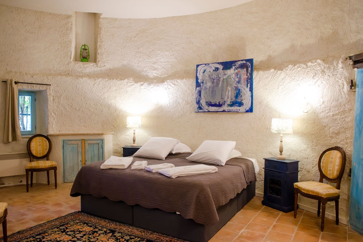 Un dormitorio con cama, dos mesitas de noche, dos lámparas, un cuadro, una ventana y una silla.