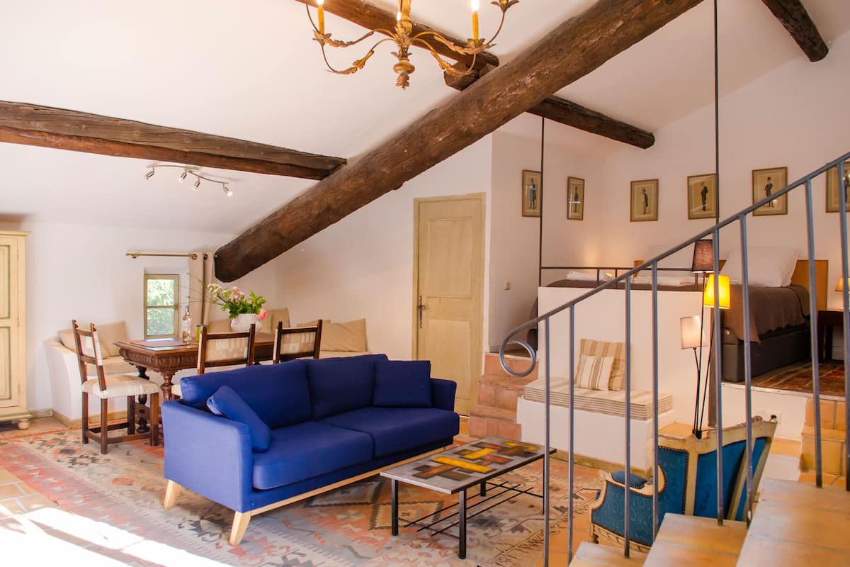 Una habitación con un sofá azul, una mesa de comedor con sillas, una araña y una escalera con barandilla.