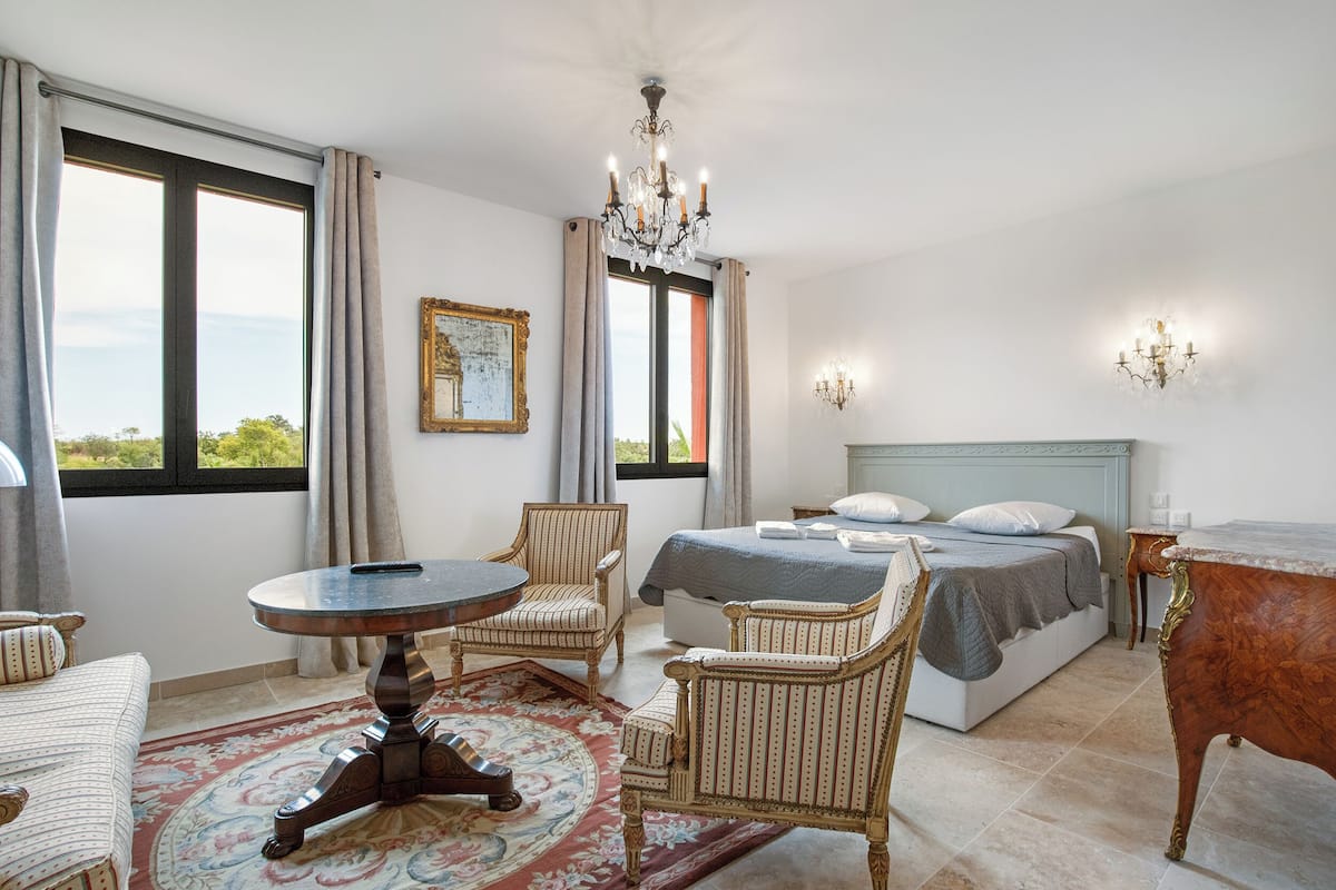 Un dormitorio con cama, dos sillones, una mesa redonda, una araña y vista al exterior.