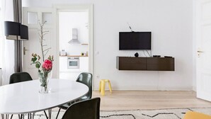 Una sala de estar moderna con una mesa de comedor, sillas, un televisor montado en la pared y una cocina visible a través de una puerta abierta.