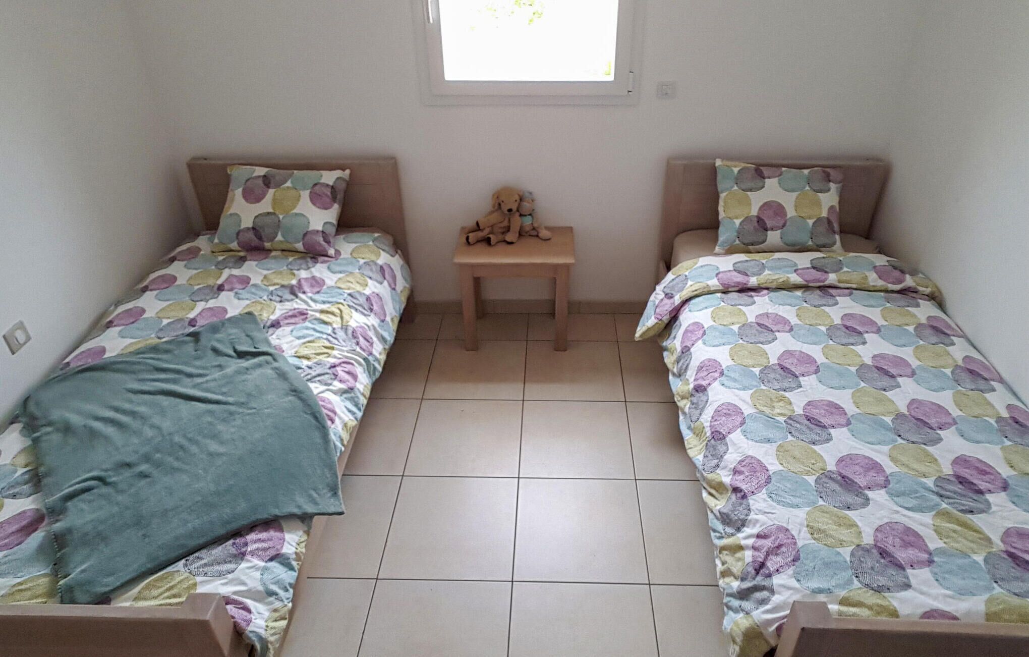 Una habitación pequeña y ordenada con dos camas individuales, cada una con una colcha y almohadas estampadas con lunares de colores. Una pequeña mesa de madera se encuentra entre las camas, y un oso de peluche está sobre ella.