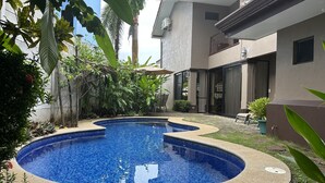Ein Schwimmbecken mit Kieselsandrand, umgeben von üppiger Bepflanzung und einem modernen Gebäude mit Balkon.