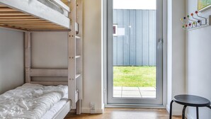 Quarto com cama beliche, um banquinho pequeno, porta que dá para um quintal e uma tomada na parede.