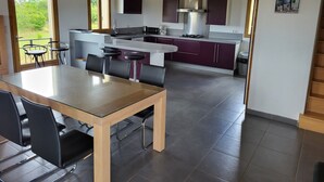 A modern kitchen with a central island, dark cabinets, and a stainless steel range hood. There is a dining area with a glass table and black chairs. - Entire villa 230 m2 with magnificent view (Blars)