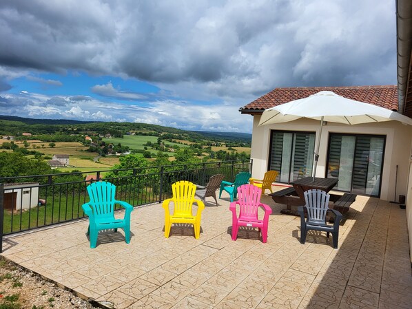 Uma varanda com cadeiras de plástico coloridas, uma mesa e um guarda-sol branco.