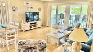 Een woonkamer met een televisie, houten meubels, een plafondventilator en uitzicht op de omgeving door grote ramen.