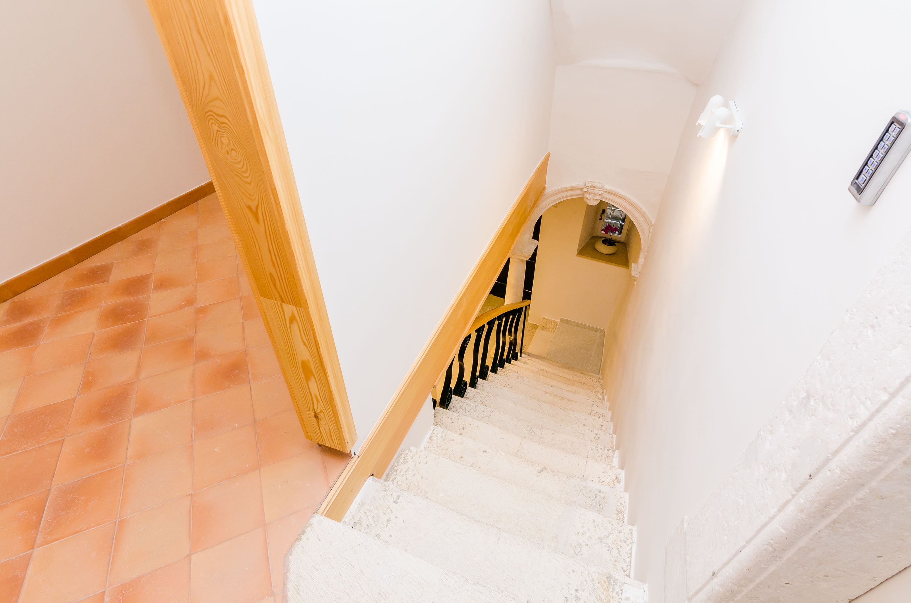 Eine Treppe mit weißen Stufen und einem hölzernen Geländer, die zu einem Zwischenpodest mit einer weißen Wand und einer Leuchte führt.