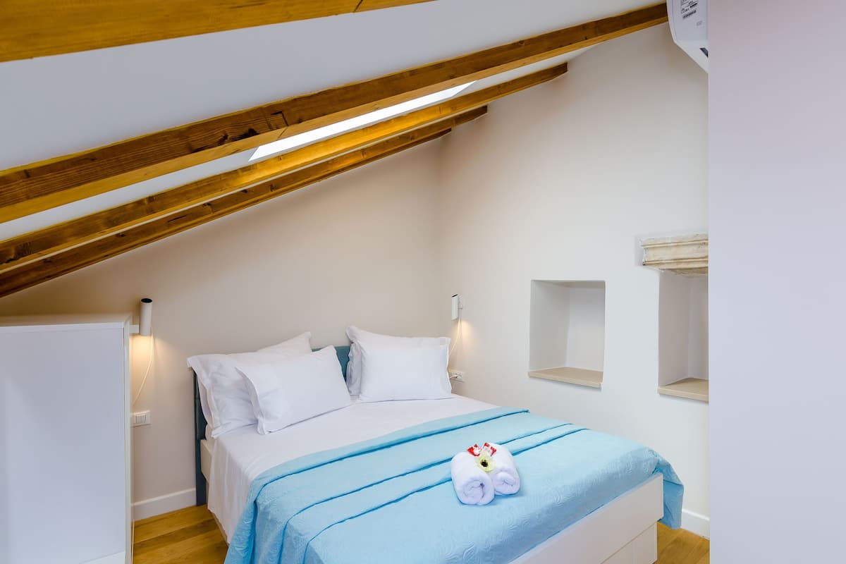 Ein Schlafzimmer mit schräg stehender Decke, einem Bett mit weißer Bettwäsche, einer blauen Decke und einem aufgerollten Handtuch auf dem Bett.