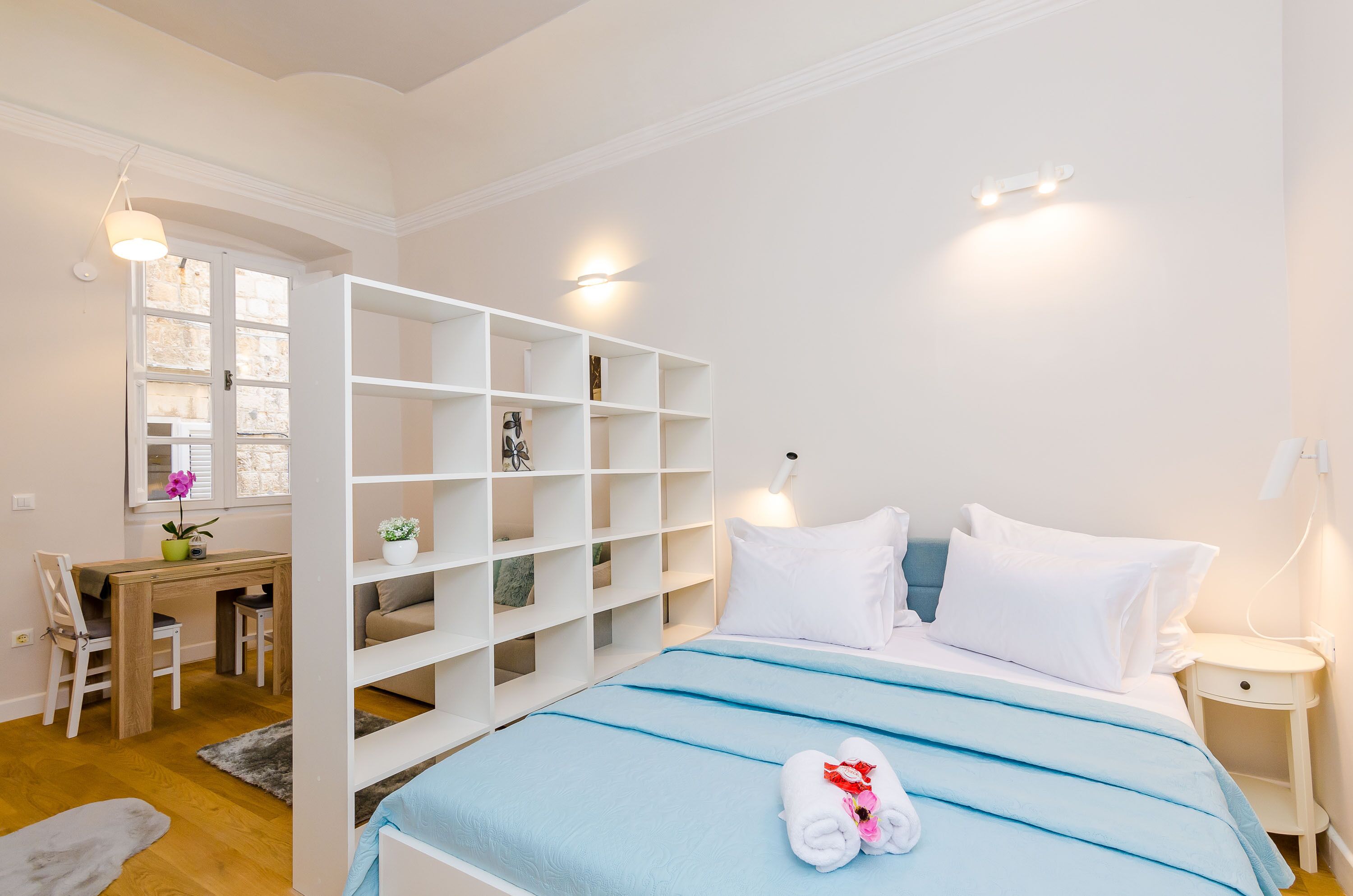 Een slaapkamer met een bed, een bureau en een stoel. Er staan planken en een nachtkastje.