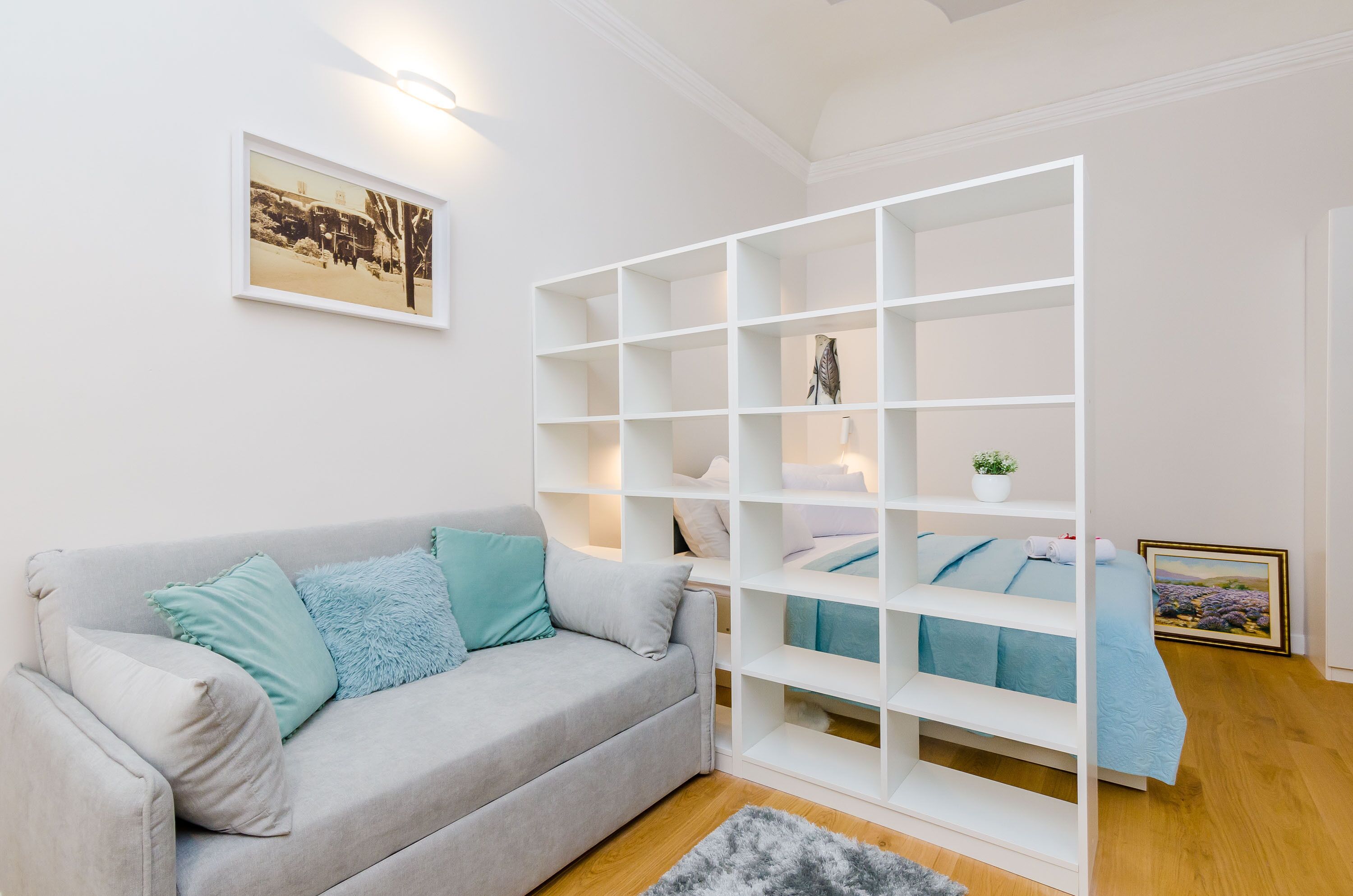 A modern living room with a grey sofa, teal cushions, a white shelving unit, and a framed picture on the wall.