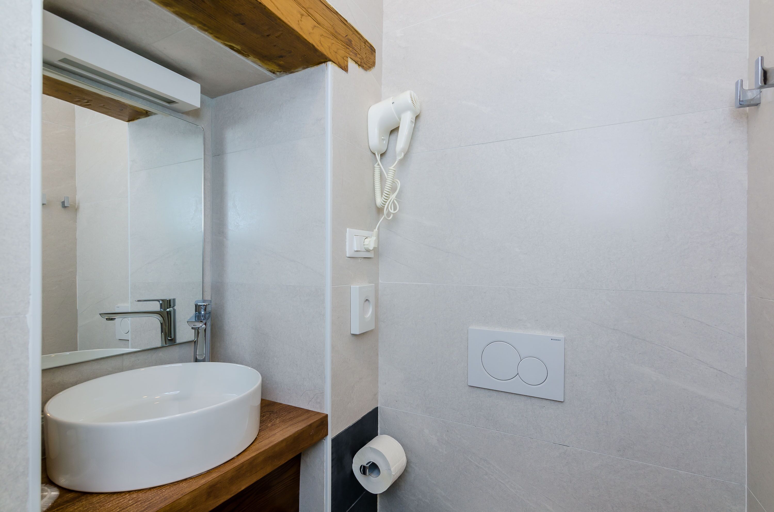 Un baño con lavamanos blanco, un espejo, un secador de pelo montado en la pared y un dispensador de papel higiénico.