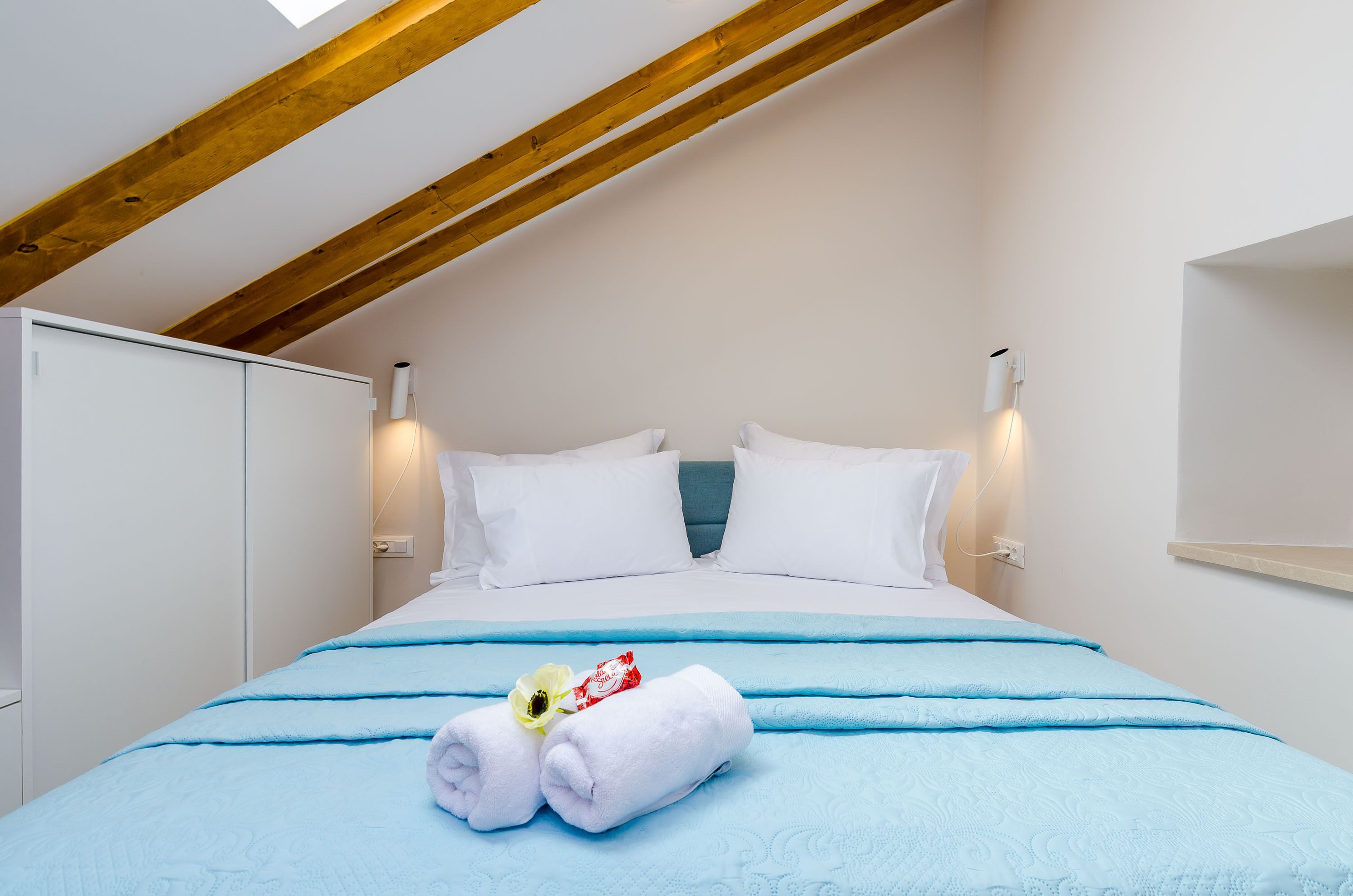 Un dormitorio con techo inclinado, paredes blancas, una cama con ropa de cama azul y dos lámparas de noche.