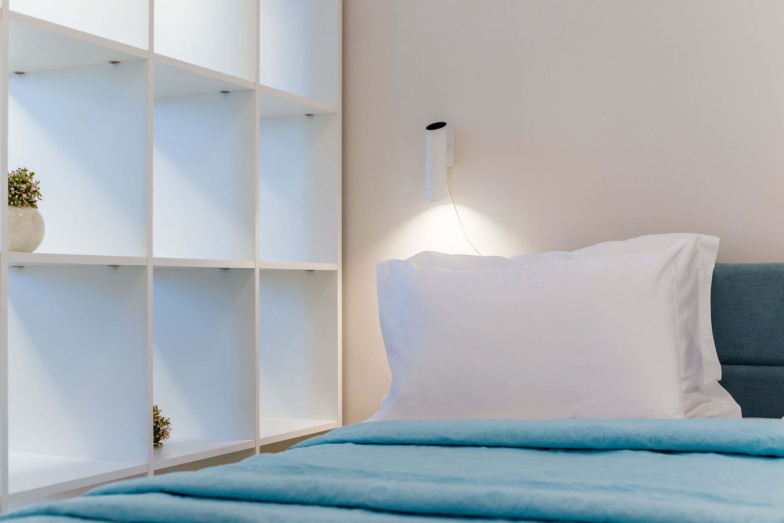 Un dormitorio con una cama, almohadas blancas, una manta azul y una lámpara blanca fijada a la pared.