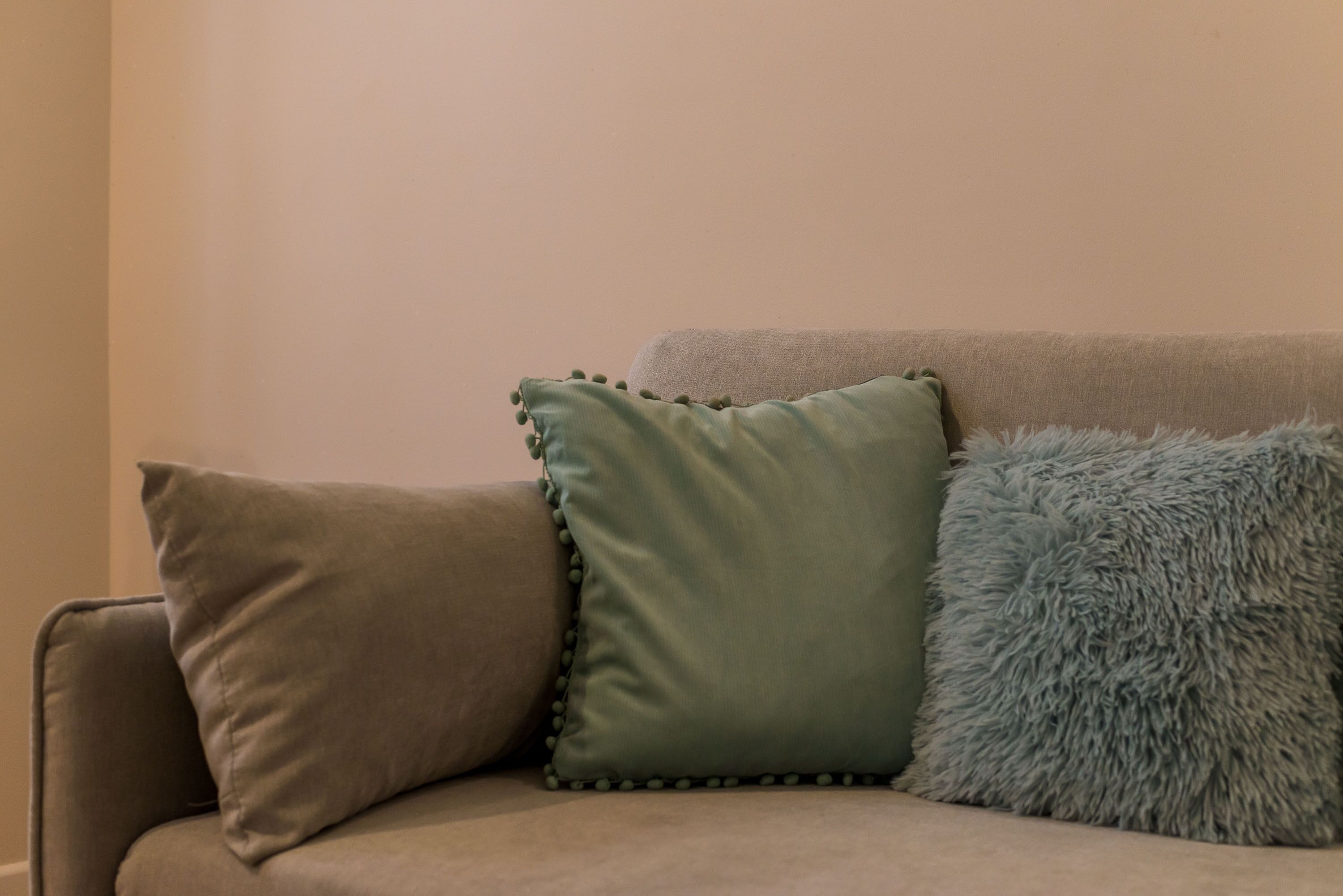 A beige sofa with three cushions: a green one with a scalloped edge, a brown one, and a fluffy blue one.
