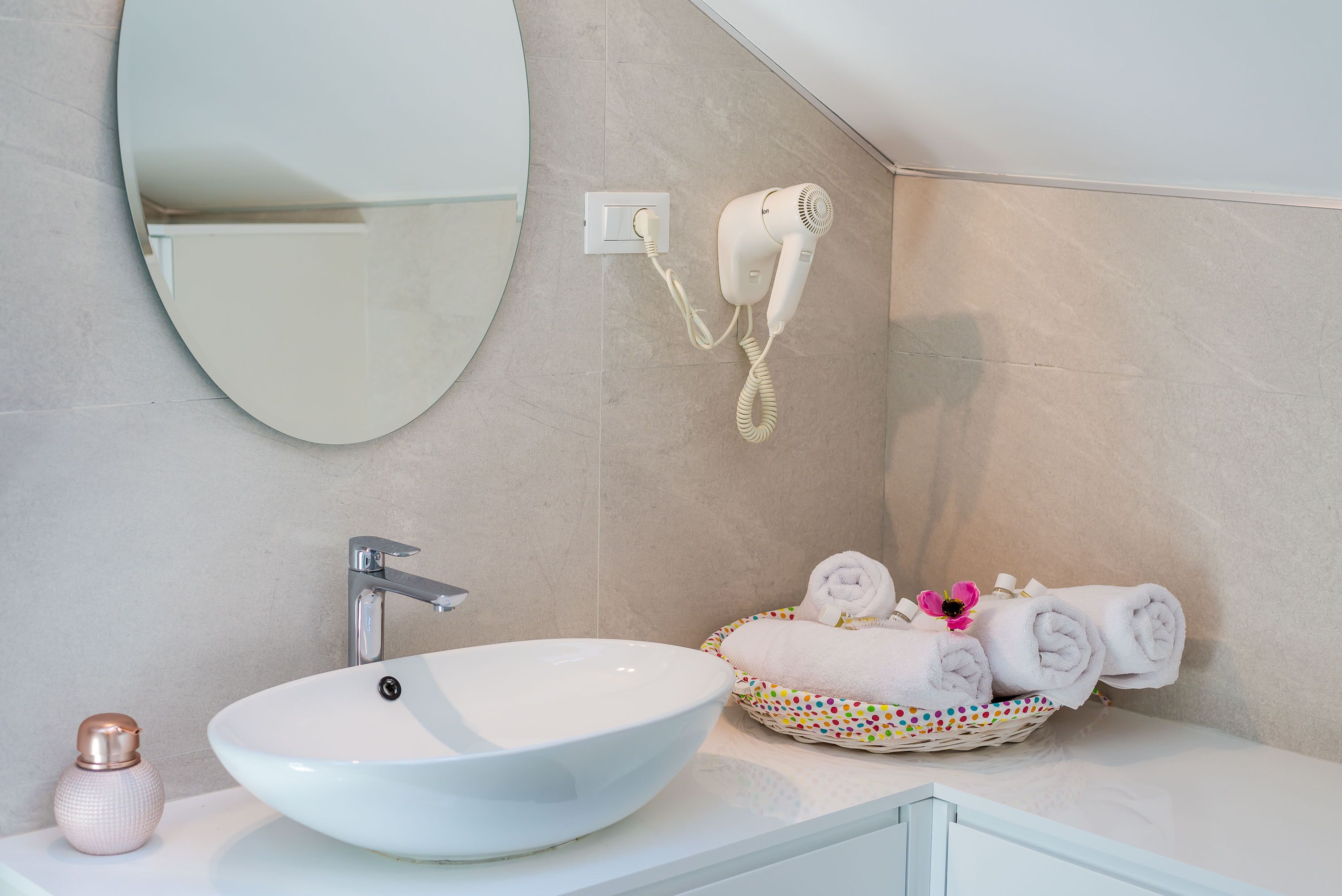 A bathroom with a white sink, a round mirror, a hairdryer mounted on the wall, and a basket of neatly folded towels.