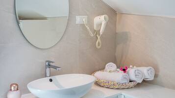 A bathroom with a white sink, a round mirror, a hairdryer mounted on the wall, and a basket of neatly folded towels.