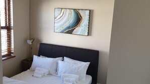 Un lit bien fait, avec du linge de lit blanc et des oreillers, une tête de lit sombre et un tableau abstrait accroché au mur.