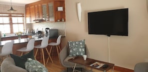 Una cocina moderna con armarios de madera, una zona de comedor con sillas blancas, un televisor de pantalla plana en la pared y un rincón de estar acogedor con cojines estampados.