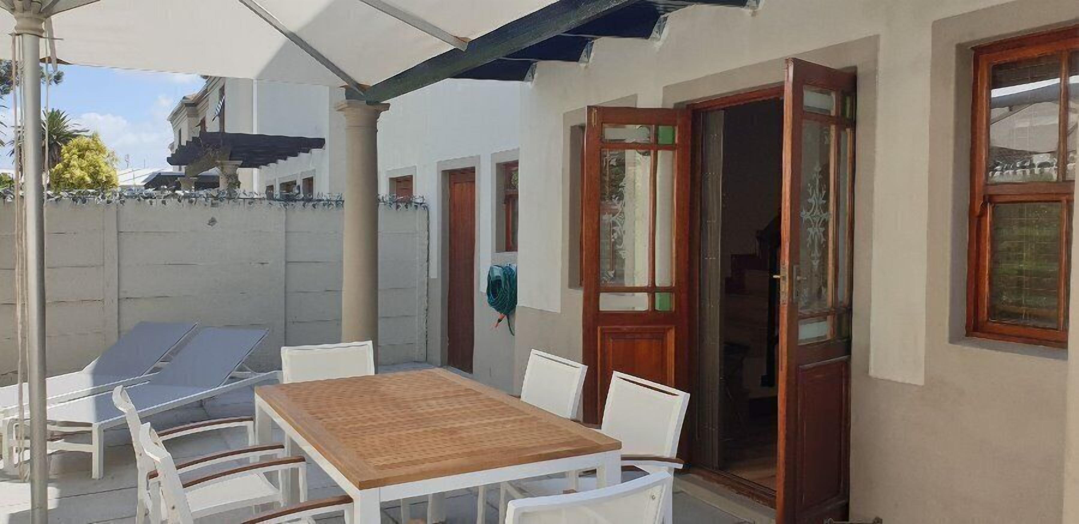 A covered outdoor seating area with a wooden table and white chairs, a wooden door, and a white wall with a window.