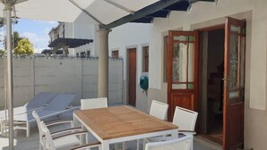 Un espace extérieur abrité, avec une table en bois et des chaises blanches, une porte en bois et un mur blanc agrémenté d’une fenêtre.