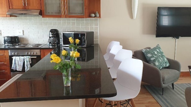 Une cuisine moderne avec un îlot central, un coin repas avec des chaises blanches, un canapé agrémenté de coussins à motifs et un téléviseur à écran plat fixé au mur.