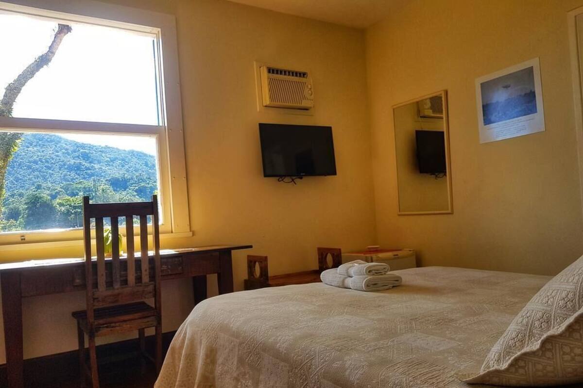 Quarto com cama, escrivaninha, cadeira, televisão e janela com vista para uma floresta.