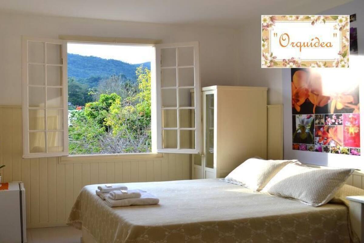 Quarto com cama, janela com venezianas abertas, pequena mesa com planta e televisão na parede.