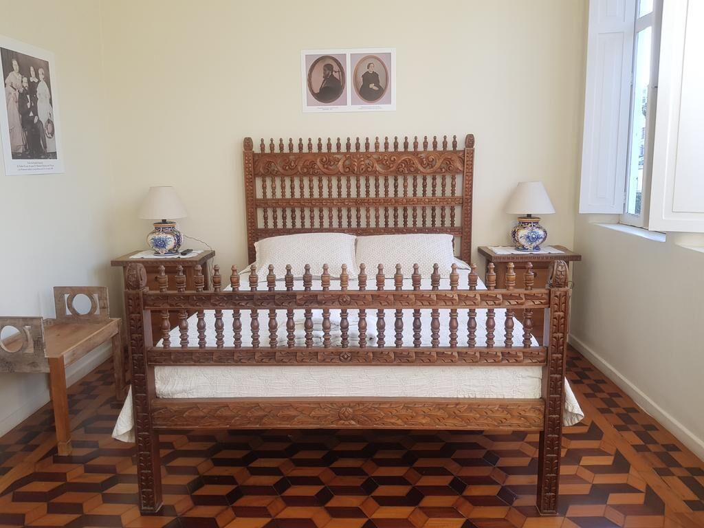 Une chambre avec un lit en bois, deux tables de chevet avec des lampes, un banc en bois, un parquet à motifs et deux tableaux encadrés accrochés au mur.