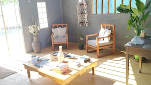 A room with a wooden coffee table, two chairs, a small table with books, a vase with dried flowers, and a hanging macrame wall decor. - Guapuruvu Suítes & Camping (Paraty)