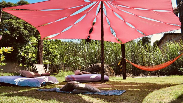 A hammock under a red shade sail in a garden with a picnic table and pillows. - Guapuruvu Suítes & Camping (Paraty)