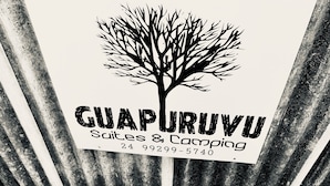 A sign for Guaparuvu Suites & Camping with a tree logo, stating the name and a contact number. - Guapuruvu Suítes & Camping (Paraty)