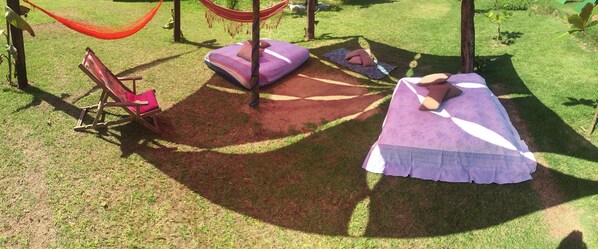 A hammock, a wooden chair, and a bed with a purple blanket and pillows in a grassy outdoor area.