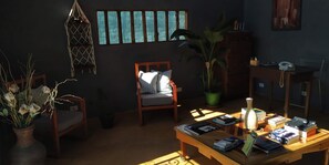 A cozy living room with a wooden coffee table, a comfortable chair, a large potted plant, and a bookshelf with books and a telephone.