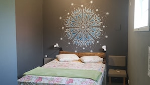 A bedroom with a bed, two bedside lamps, a floral bedspread, a green throw blanket, a wall with a geometric design, a window, and a chair. - Guapuruvu Suítes & Camping (Paraty)