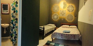 A bedroom with a bed, a bedside table, a nightstand, a wall-mounted light fixture, and a bathroom with a sink and a mirror.