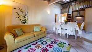 Una sala de estar con un sofá, un tapete colorido, un comedor con mesa y sillas, y una cocina al fondo.