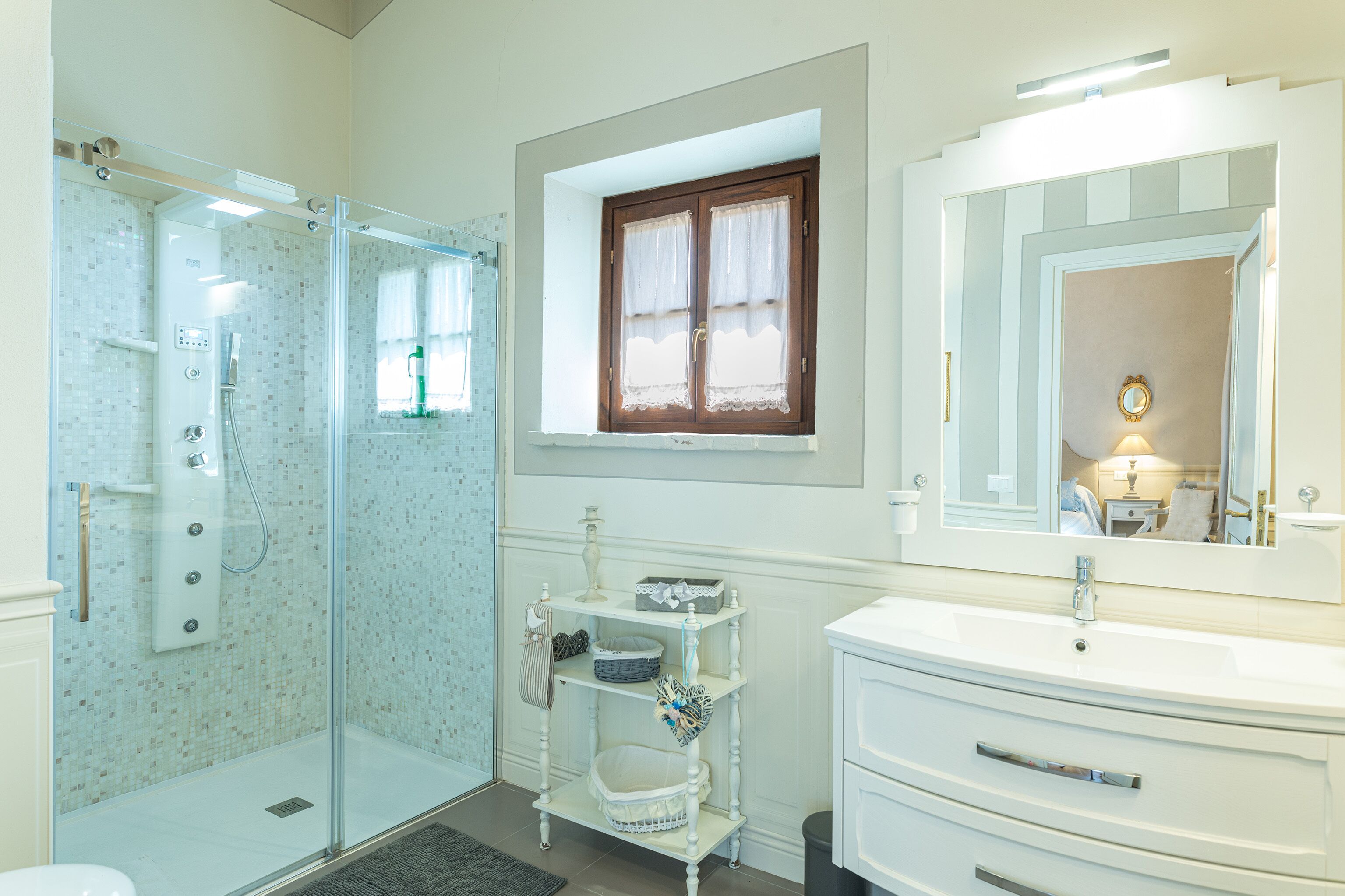A bathroom with a glass-enclosed shower, a vanity with a mirror, and a small window with curtains.