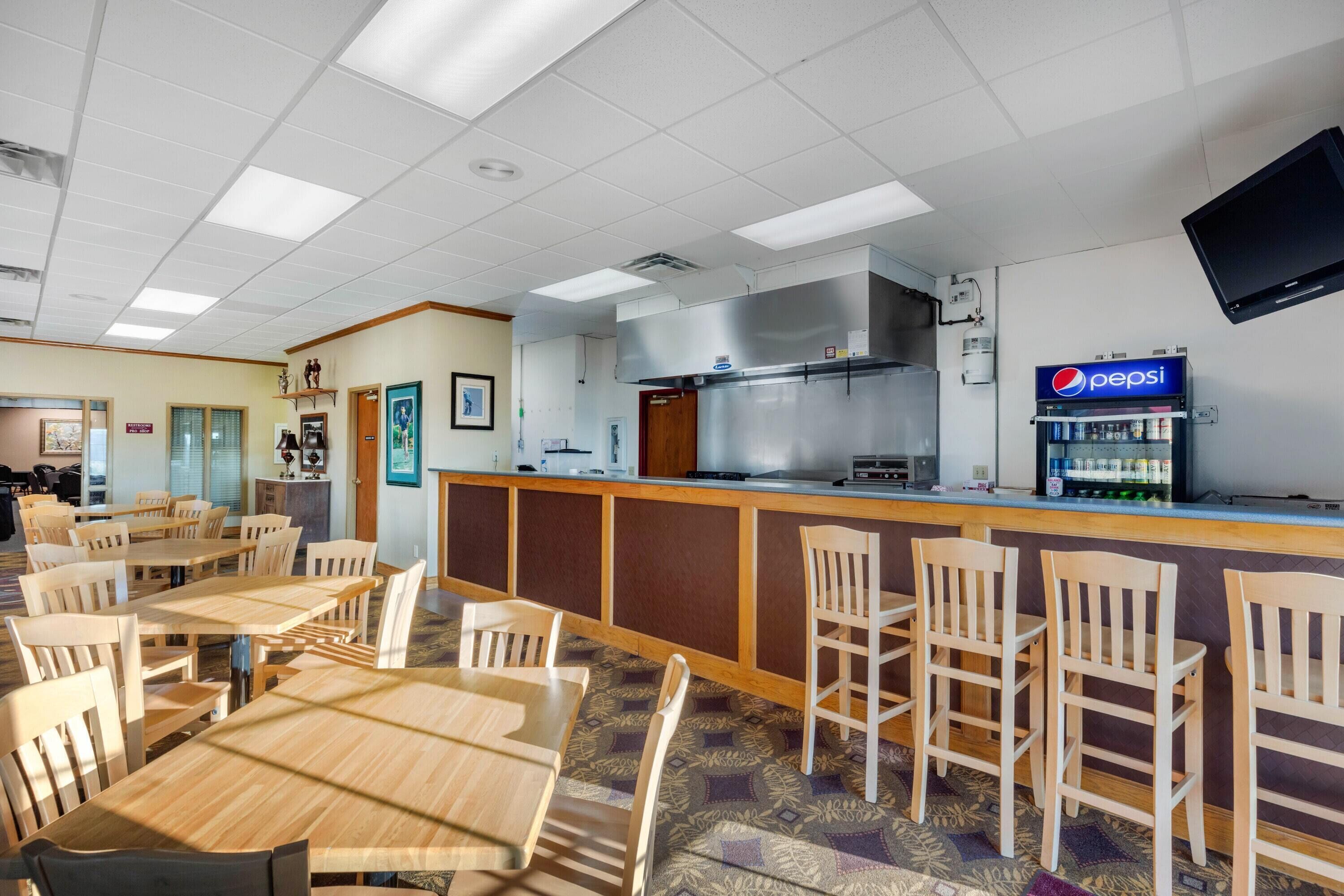Un restaurante con mesas y sillas de madera, una zona de bar con refrigerador de Pepsi y una cocina al fondo.