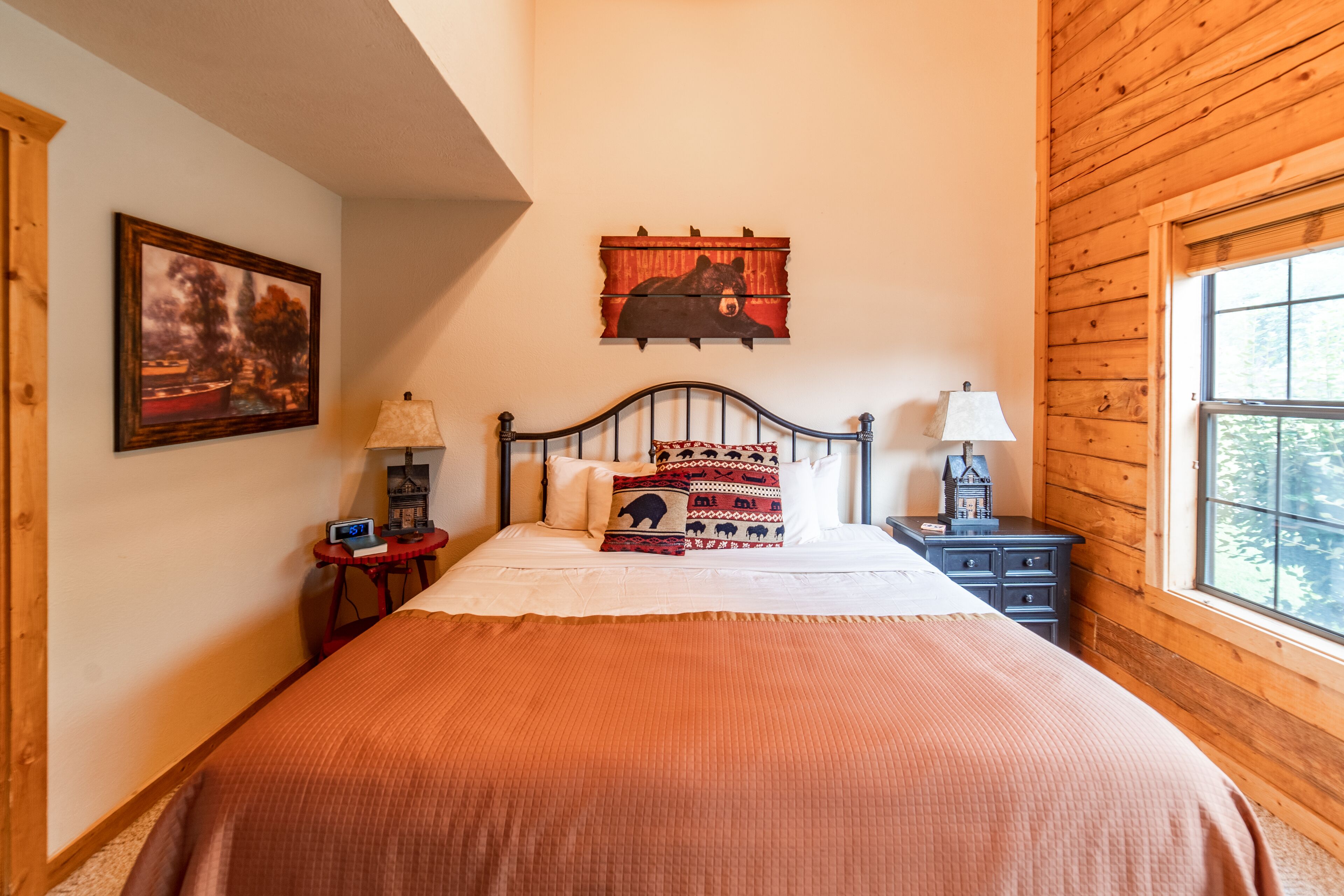 A bedroom with a wooden ceiling, a bed with a patterned blanket, two bedside tables with lamps, a wall-mounted clock, and a framed picture.