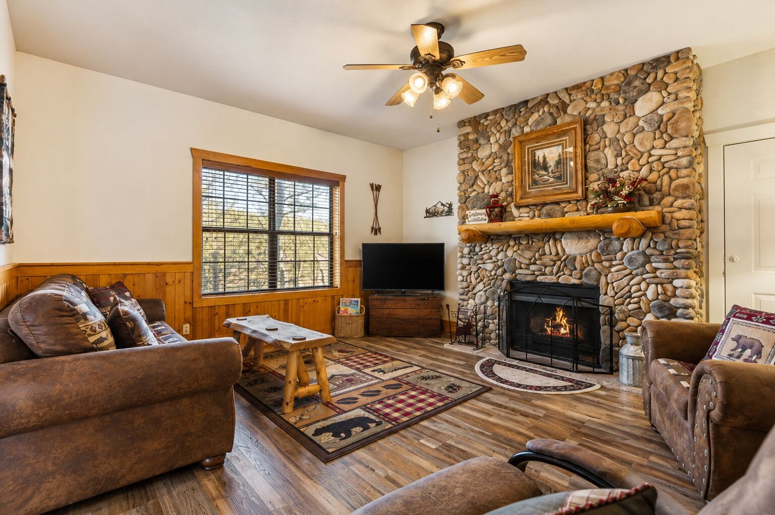 A cozy living room with a stone fireplace, wooden furniture, and a large window.