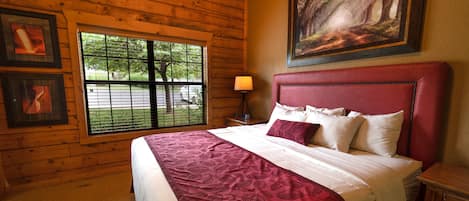 A bedroom with a large bed, a window with blinds, a nightstand, and a painting on the wall.