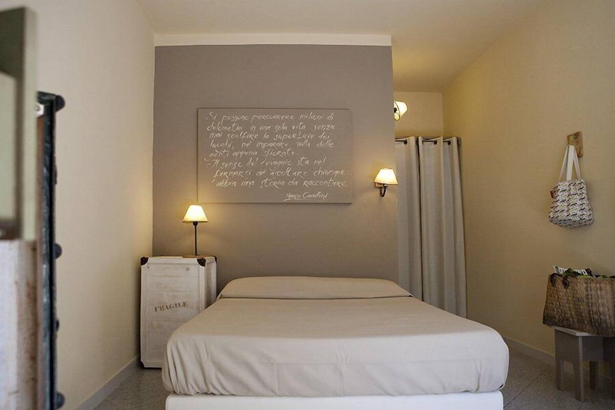 Une chambre d’hôtel avec un lit, une citation affichée sur le mur, deux lampes murales, un radiateur, une valise et une petite table sur laquelle se trouve un sac.